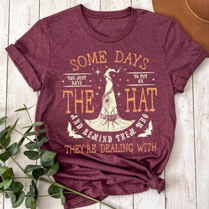 May include: Burgundy t-shirt with the text "SOME DAYS YOU JUST HAVE TO PUT ON THE HAT AND REMIND THEM WHO THEY'RE DEALING WITH." The graphic features a witch's hat with a spider and bats. The shirt is knotted at the bottom.