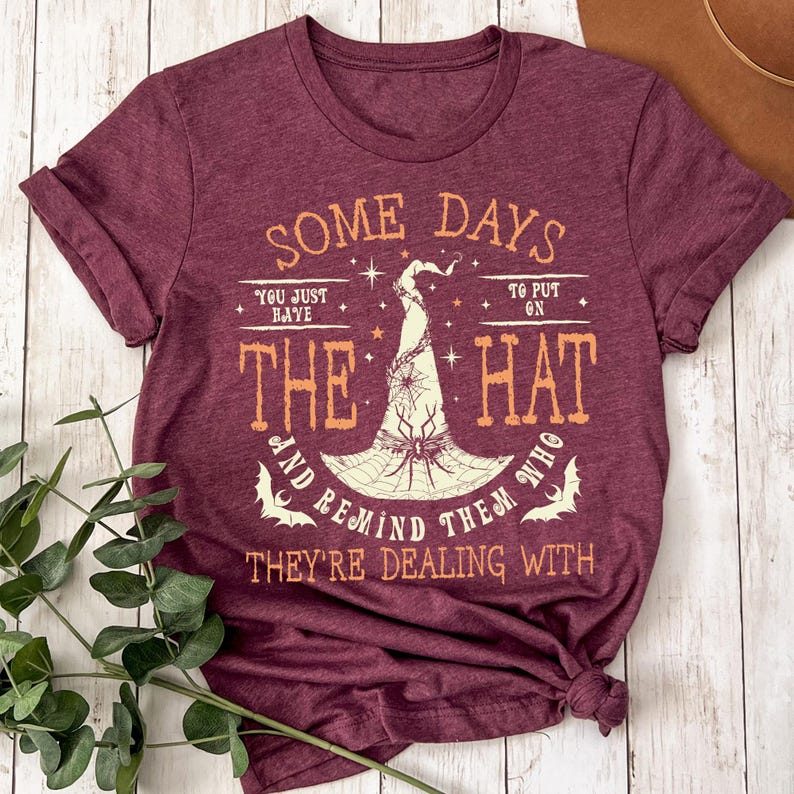 May include: Burgundy t-shirt with the text "SOME DAYS YOU JUST HAVE TO PUT ON THE HAT AND REMIND THEM WHO THEY'RE DEALING WITH." The graphic features a witch's hat with a spider and bats. The shirt is knotted at the bottom.