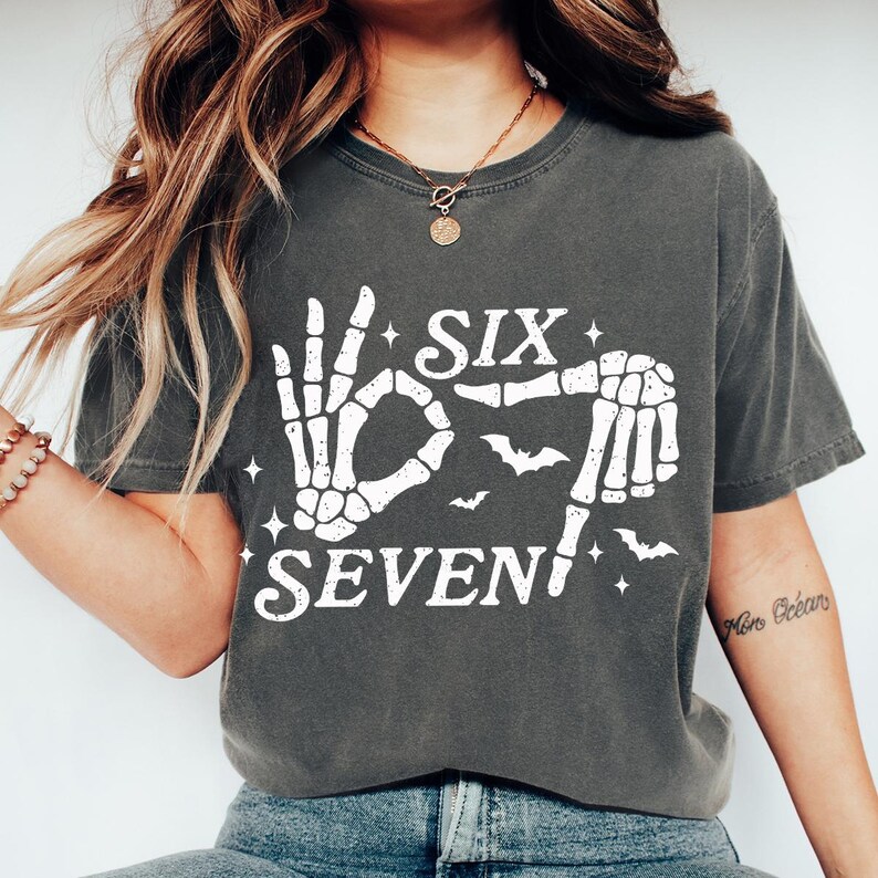 May include: Dark gray t-shirt with a white skeleton hand graphic forming the number six and seven. The design includes bats and stars. The shirt is a casual, unisex style.