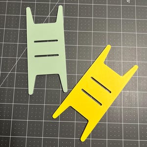 May include: Two plastic craft tools, one green and one yellow, shaped like ladders with three rungs. The tools are on a black grid surface.