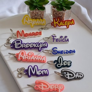 Op de afbeelding: Een verzameling kleurrijke naam sleutelhangers met namen als Arpana, Kalea en Monroe. Elke sleutelhanger heeft een unieke kleurencombinatie en een zilveren sleutelring. De sleutelhangers worden gepresenteerd op een witte ondergrond.