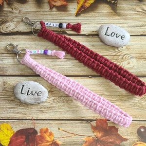 May include: Two macrame wrist lanyards in pink and burgundy, each with a silver clasp and beaded name tag. The pink lanyard has the name "NORI" and the burgundy lanyard has the name "MAII". A stone with the word "Love" and another with "Live" are also in the image.