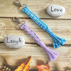 Könnte beinhalten: Zwei Makramee-Schlüsselanhänger, einer blau und einer lila, jeweils mit einem Namen, der in Buchstabenperlen geschrieben ist. Der blaue Schlüsselanhänger sagt "MIKE" und der lila Schlüsselanhänger sagt "RUBY". Die Schlüsselanhänger haben silberne Verschlüsse und Quasten. Das Wort "Love" steht auf einem Stein.