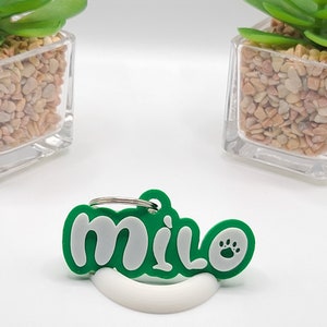May include: A green and white pet tag with the name "milo" and a paw print. The tag is made of a rubbery material and has a silver keyring attached. The tag is resting on a white curved stand.