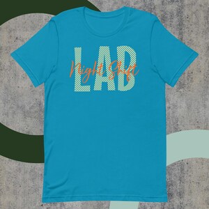 Könnte beinhalten: Ein türkisfarbenes T-Shirt mit dem Wort "LAB" in großen weißen gepunkteten Buchstaben und den Worten "Night Shift" in orangefarbener Schrift. Das Shirt besteht aus einem weichen Material und hat kurze Ärmel.