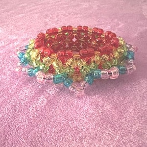 Può includere: Un braccialetto di perline multicolore. Il braccialetto presenta una sfumatura di colori, tra cui rosso, arancione, giallo, verde, blu e rosa. Le perline sono piccole e quadrate, creando un aspetto strutturato. Il braccialetto è su uno sfondo rosa.