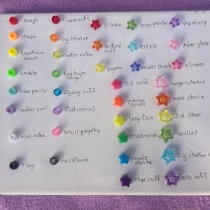 Pu&ograve; includere: Una tela bianca mostra un assortimento di perline colorate e abbellimenti a forma di stella. Etichette scritte a mano identificano ogni articolo, tra cui "single", "flower cuff", "X-base" e "mystery". Le perline sono in vari colori come rosso, arancione, giallo, verde, blu e viola.