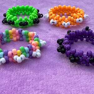Può includere: Quattro braccialetti di perline colorate in verde, arancione, arcobaleno e viola. I braccialetti sono fatti di piccole perline rotonde e sono disposti su una superficie viola. I braccialetti verdi e arancioni hanno accenti neri e bianchi.