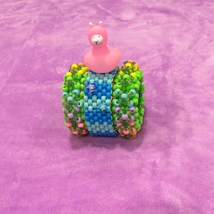 Può includere: Un braccialetto di perline colorate con una statuina di lama rosa sopra. Il braccialetto ha un design a cascata con perline blu, verdi e gialle. Lo sfondo è di colore viola.