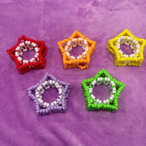Può includere: Cinque braccialetti a forma di stella con perline rosse, arancioni, gialle, viola e verdi. Ogni braccialetto presenta una forma a stella con un anello interno bianco. I braccialetti sono fatti di piccole perline colorate.