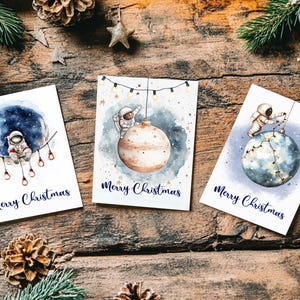 May include: Three Christmas cards with watercolour illustrations of astronauts decorating planets. Each card features the text "Merry Christmas" in a dark blue script. The cards are arranged on a rustic wooden surface with pine cones and greenery.