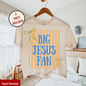Può includere: T-shirt beige con la scritta blu "BIG JESUS FAN" su uno sfondo a righe verticali arancioni. La maglietta presenta fiocchi gialli ed è appesa a una gruccia di legno.