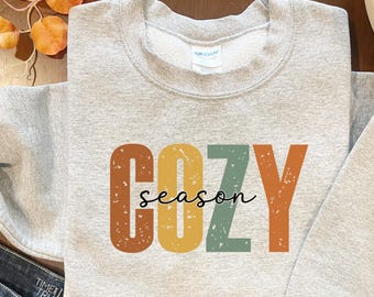 May include: A light gray sweatshirt with the words "COZY SEASON" printed on the front. The word "COZY" is in large, distressed, block letters in shades of brown, yellow, and green. The word "season" is in a cursive font.