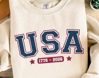 Può includere: Felpa color crema con la scritta "USA" in lettere blu navy con contorno rosso. L'intervallo di anni "1776 - 2026" è sotto le lettere, con stelle rosse su entrambi i lati. Le parole "DOWNLOAD DIGITALE" sono in alto.