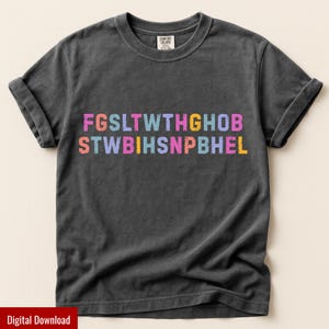 Könnte beinhalten: Dunkelgraues T-Shirt mit hochgekrempelten Ärmeln. Das Shirt zeigt farbigen Text in horizontaler Anordnung: "FGSLT WTHGHOB" und "STWB IHSNPBHEL". Das Shirt ist aus weichem Material und hat einen klassischen Rundhalsausschnitt.