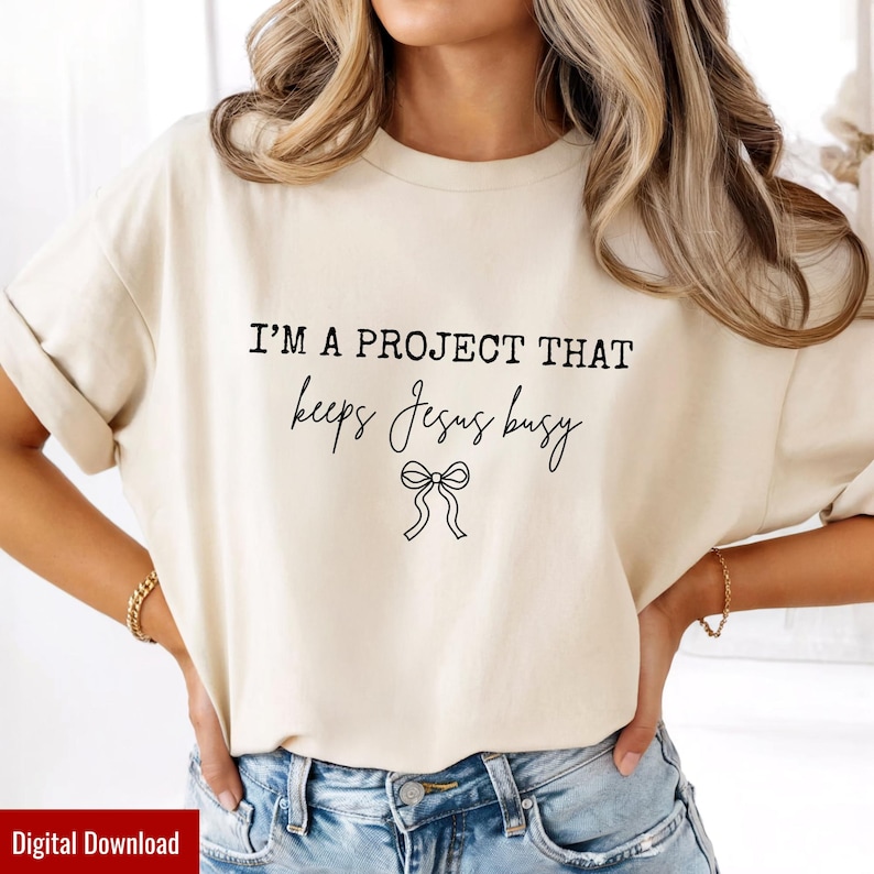 May include: Cream-colored t-shirt with the text "I'M A PROJECT THAT keeps Jesus busy" in black script. A bow graphic is below the text. The shirt is a casual style.