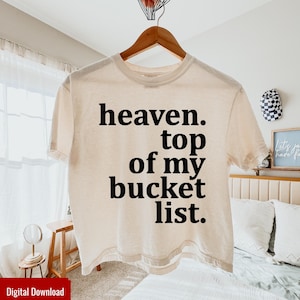 May include: Cream-colored t-shirt with the text "heaven. top of my bucket list." in black. The shirt is hanging on a wooden hanger. The background includes a bed, a small table, and a decorative item.