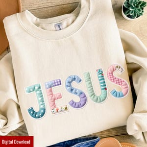 May include: A cream-coloured sweatshirt featuring the word "JESUS" appliquéd across the front. The letters are crafted from various patterned fabrics in pastel shades, such as polka dots, gingham, and floral designs. The sweatshirt is a relaxed, comfortable style.