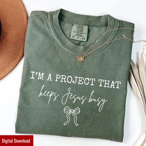May include: Olive green Comfort Colors t-shirt with the text "I'M A PROJECT THAT keeps Jesus busy" in white script. A gold necklace with a pendant rests on the shirt. A brown hat is visible in the background.