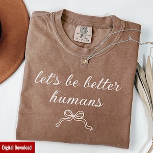May include: A brown Comfort Colors t-shirt with the phrase "let's be better humans" in white script. A silver necklace with a small pendant rests on the shirt. The shirt has a small bow graphic below the text.