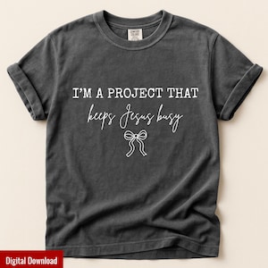 May include: Dark gray t-shirt with rolled-up sleeves. The shirt features the white text "I'M A PROJECT THAT keeps Jesus busy" with a bow graphic. The shirt is made of a soft material and is a Comfort Colors brand.