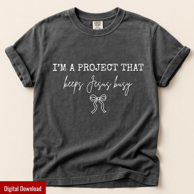 May include: Dark gray t-shirt with rolled-up sleeves. The shirt features the white text "I'M A PROJECT THAT keeps Jesus busy" with a bow graphic. The shirt is made of a soft material and is a Comfort Colors brand.
