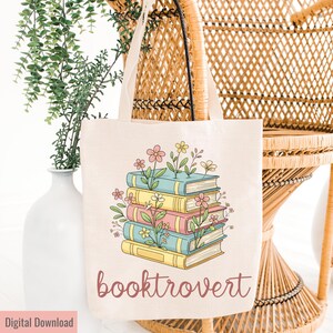 May include: A beige tote bag featuring a stack of colorful books with flowers growing from them. The text "booktrovert" is printed in a cursive font below the books. The bag is hanging on a wicker chair.