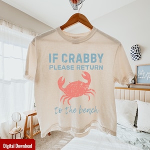 May include: Cream-colored t-shirt with the text "IF CRABBY PLEASE RETURN to the beach" in light blue. A coral-colored crab graphic is centered below the text. The shirt is hanging on a wooden hanger.