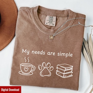 May include: Brown Comfort Colors t-shirt with the phrase "My needs are simple" and illustrations of a coffee cup, paw print, and books. A silver necklace with a pendant is draped across the shirt. The shirt is a size small.