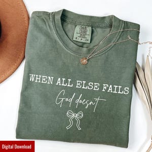 Può includere: Una maglietta verde oliva con la scritta bianca "WHEN ALL ELSE FAILS God doesn't". Una collana d'oro con un ciondolo è appoggiata sulla maglietta. La maglietta ha una piccola etichetta con la scritta "COMFORT COLORS".