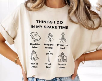 May include: Cream-colored t-shirt with the text "THINGS I DO IN MY SPARE TIME" and illustrations of a bible, praying hands, a cross, and a church. The shirt also includes the phrases "Read the bible", "Pray the rosary", "Praise the lord", "Talk to Jesus", "Trust God", and "Grow in faith".