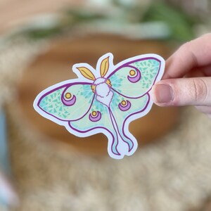 May include: A sticker of a green and purple luna moth with yellow accents. The moth has a detailed pattern of small circles and lines.