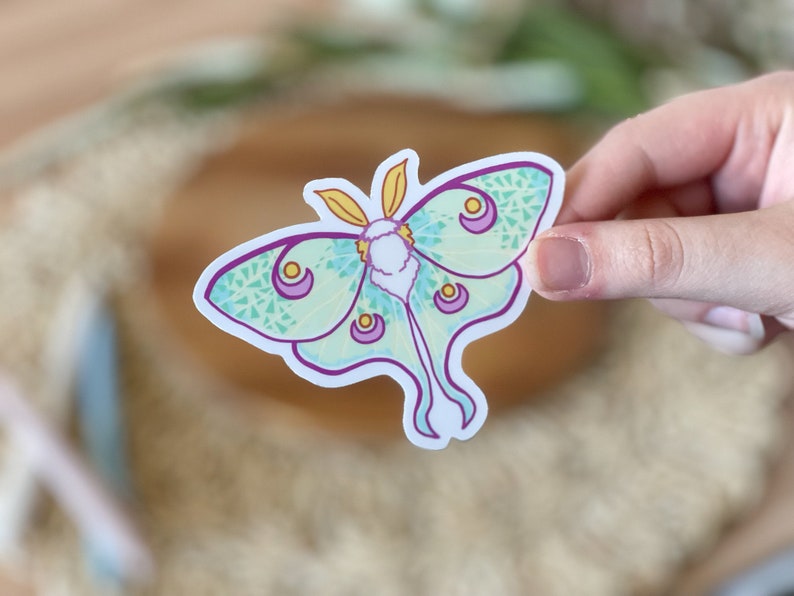 May include: A sticker of a green and purple luna moth with yellow accents. The moth has a detailed pattern of small circles and lines.