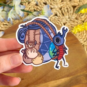 May include: A sticker featuring a cartoon illustration of a backpack and a beetle. The backpack is brown and blue with a rope handle and two small pouches. The beetle is blue and red with large eyes and a curved horn.