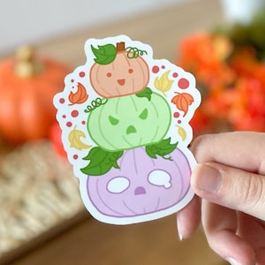May include: A sticker of three stacked pumpkins with different facial expressions. The top pumpkin is orange with a happy face, the middle pumpkin is green with an angry face, and the bottom pumpkin is purple with a sad face. The pumpkins are surrounded by small red and orange leaves.