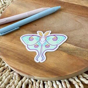 May include: A green and purple luna moth sticker with yellow accents on a wooden surface.