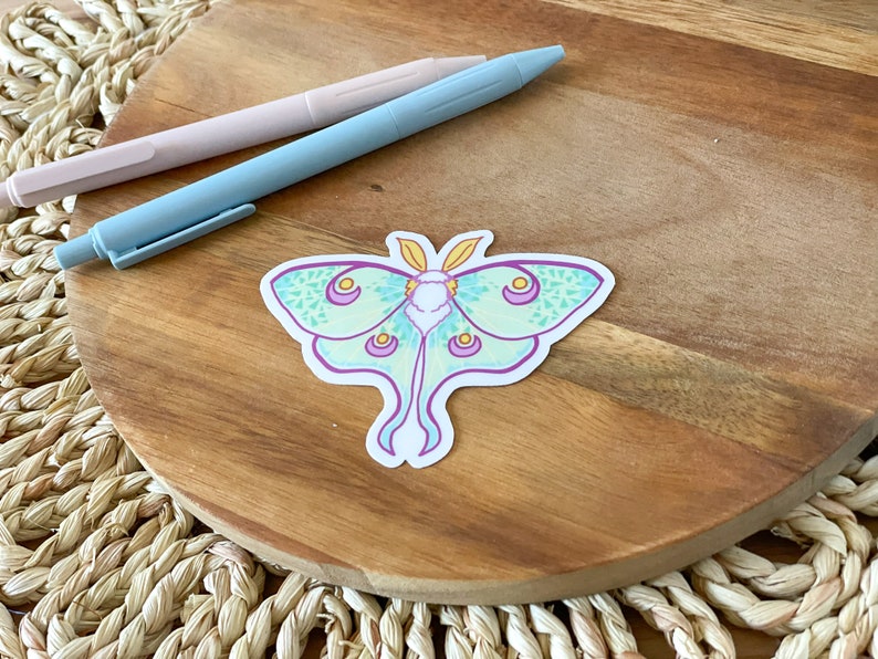 May include: A green and purple luna moth sticker with yellow accents on a wooden surface.