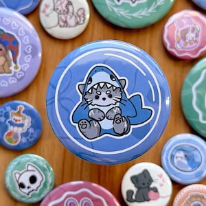 May include: A blue and white button with a cartoon cat wearing a shark costume. The cat is gray and white with a pink nose and blue eyes. The shark costume is blue with white teeth and a white fin. The button is surrounded by other buttons with various designs.