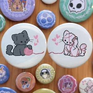 May include: Two round pins with a white background and pink hearts. One pin features a black cat holding a ball of yarn. The other pin features a white cat tangled in yarn.