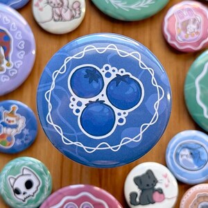 Op de afbeelding: Blauwe en witte knoop met een ontwerp van drie blauwe bessen op een blauwe achtergrond. De blauwe bessen zijn omringd door een wit kant-achtig ontwerp.