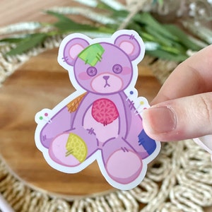 May include: A purple patchwork teddy bear with a green patch over one eye. The bear has a pink heart on its chest and is stitched together with colorful thread.