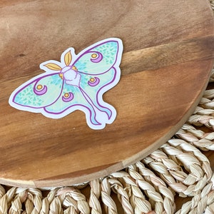 May include: A green and purple luna moth sticker with yellow accents on a wooden surface.