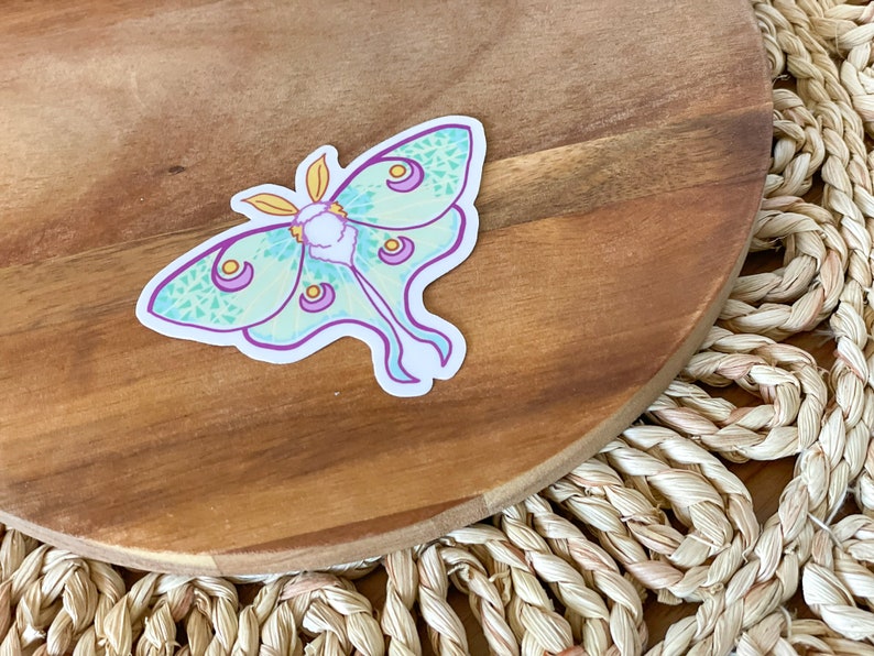 May include: A green and purple luna moth sticker with yellow accents on a wooden surface.