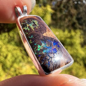 May include: A rectangular pendant with a silver frame. The pendant features a dark brown opal with iridescent blue and green flashes. The opal has a cracked, textured appearance. The pendant is held up by a silver bail.
