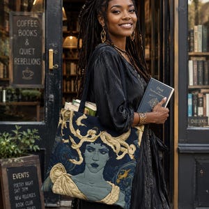 Op de afbeelding: Een donkerblauwe tote bag met een doorgestikte grafische illustratie van een figuur met goudkleurig slangachtig haar. De tas heeft twee zwarte schouderbanden en bevat diverse boeken. Het ontwerp bevat gedetailleerde gouden en blauwe patronen.