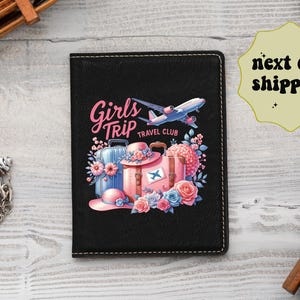 May include: A black passport holder featuring a graphic print of pink suitcases, a blue airplane, and floral accents with the text Girls Trip Travel Club. The cover has white stitching along the edges and a rectangular shape with rounded corners.