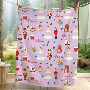 May include: A light purple blanket featuring a repeating pattern of nutcrackers, ballerinas, drums, xylophones, teddy bears, and rocking horses, interspersed with the name Olivia in red script and small green holly leaf accents.