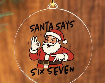 May include: A clear, round Christmas ornament with a cartoon Santa Claus illustration. The text "SANTA SAYS SIX SEVEN" is printed on the ornament. The ornament is hanging from a gold string against a wooden background.