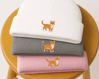 Puede incluir: Tres gorros de punto acanalado en blanco, gris y rosa, cada uno bordado con un gato atigrado naranja de dibujos animados. Los gorros están apilados en una silla de madera, mostrando las diferentes opciones de color.