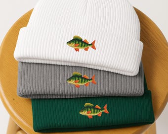 Puede incluir: Tres gorros de punto acanalado en blanco, gris y verde apilados sobre un taburete de madera. Cada gorro presenta un diseño de pez bordado en tonos verdes, amarillos y rojos. Los gorros están doblados en el borde.